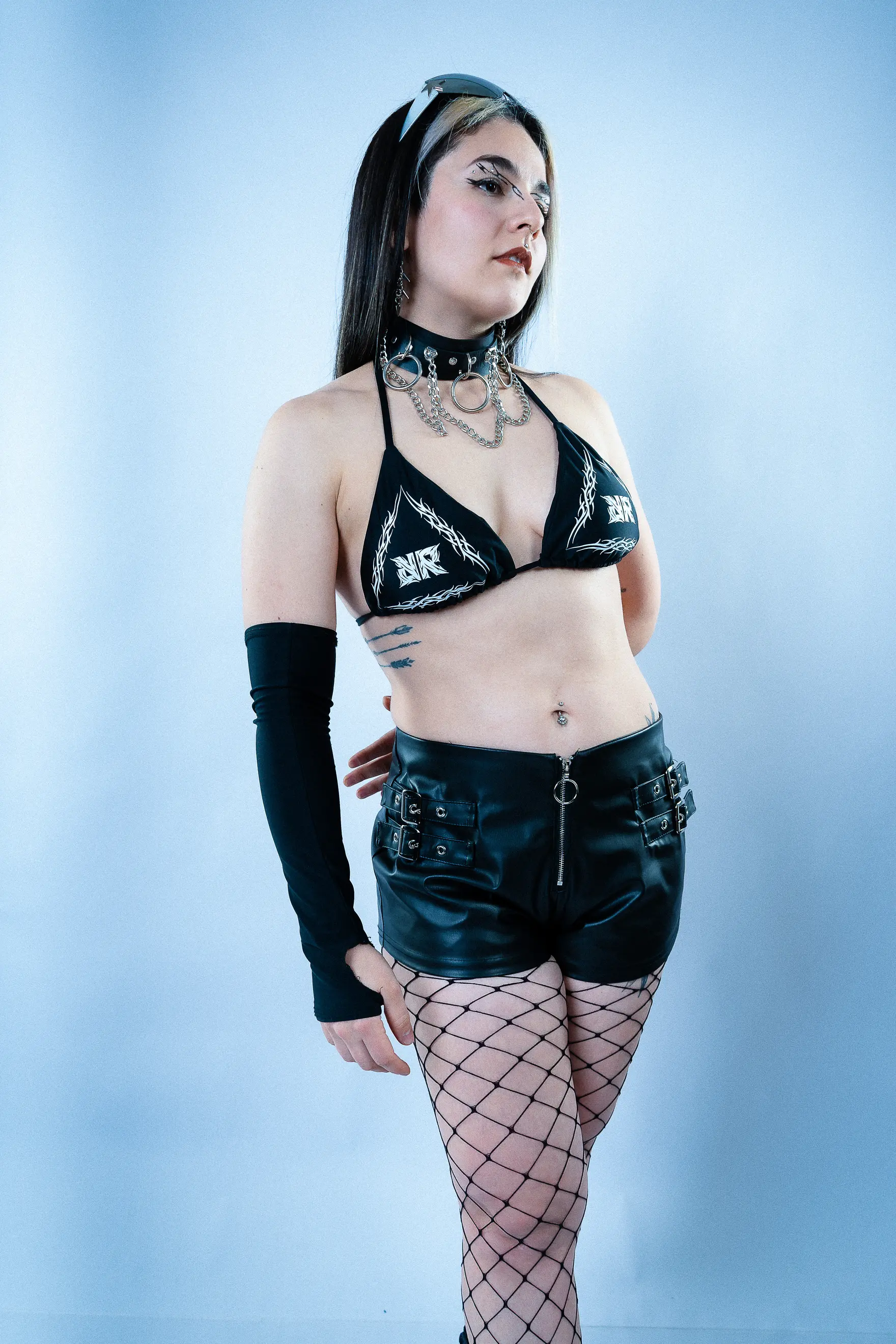 Modelo con top y shorts negros de Real Raver, guantes largos y medias de red, sobre fondo azul grisáceo de estudio.